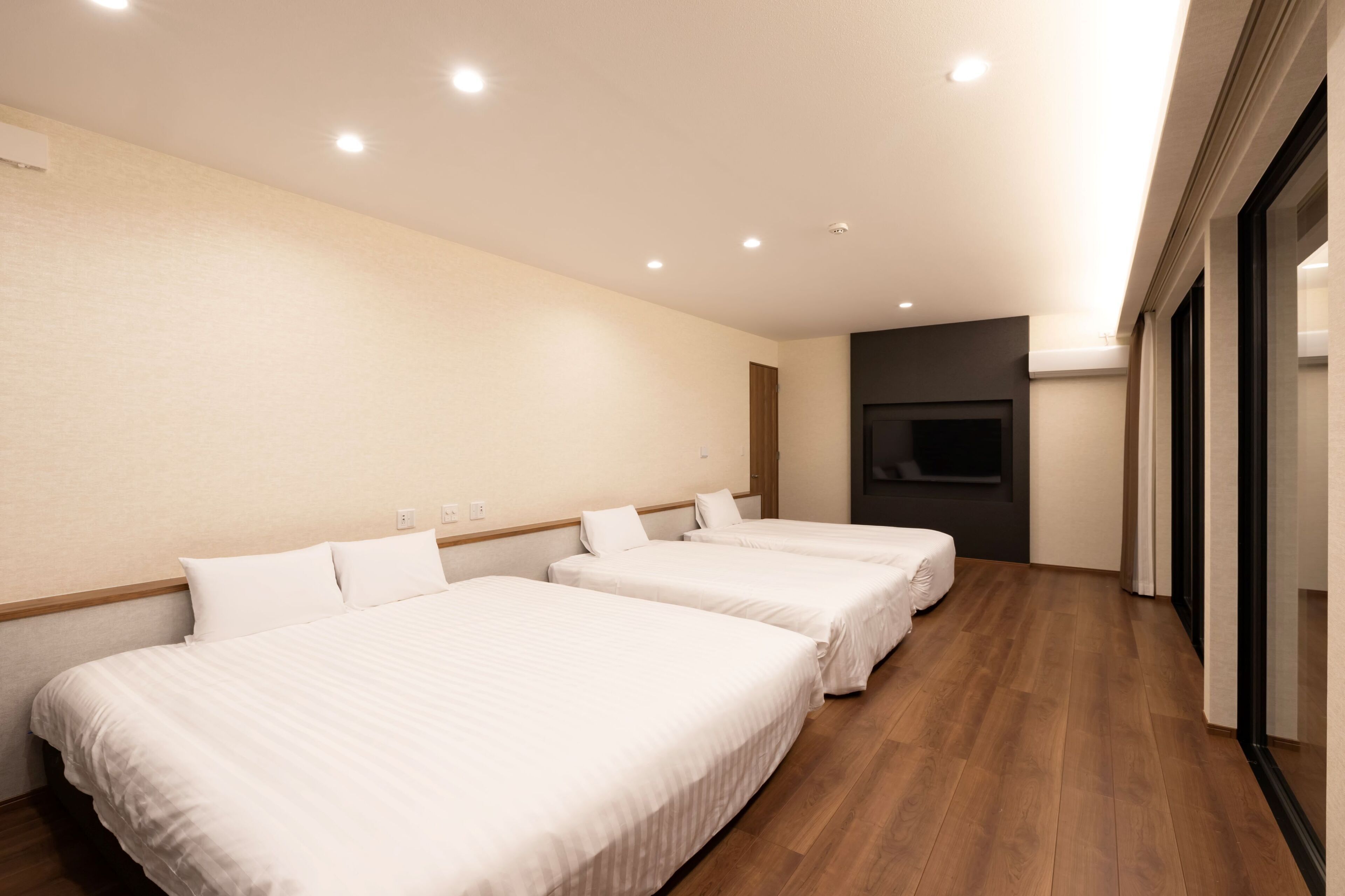 A hotel room with three beds, wooden flooring, a flat-screen TV mounted on the wall, and a large window.