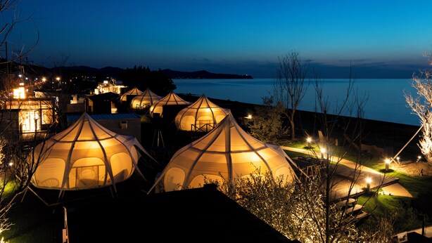A series of dome-shaped tents illuminated at dusk, with a view of a calm body of water and distant hills. - Workation Resort THE Forest (Nagatoro)