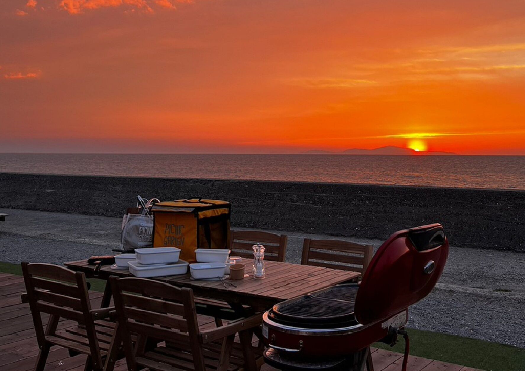 A wooden deck with a table set for a meal, a grill, and picnic supplies against a sunset view.