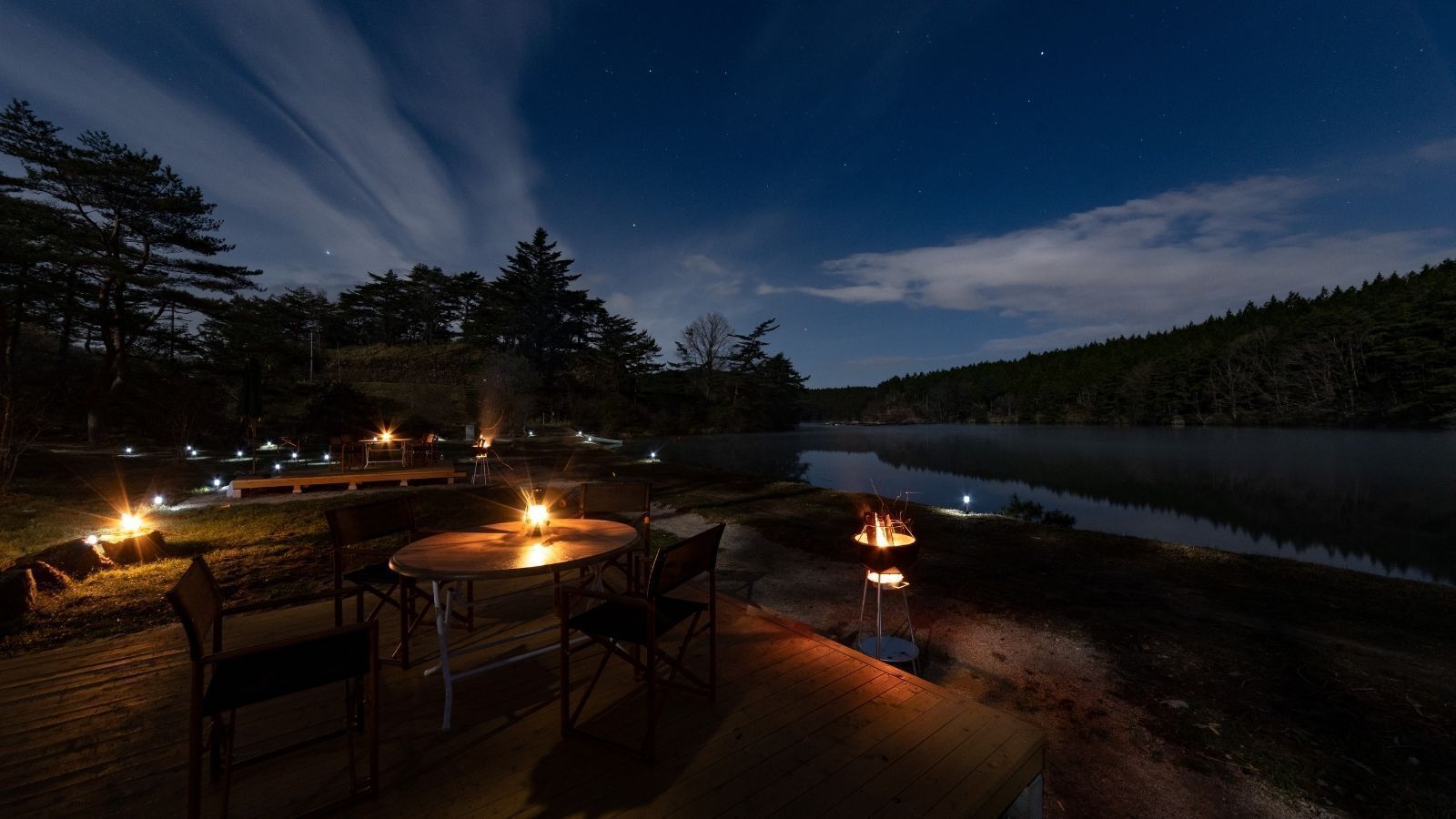 An outdoor deck with a round table and chairs, a grill, and a view of a lake at night.