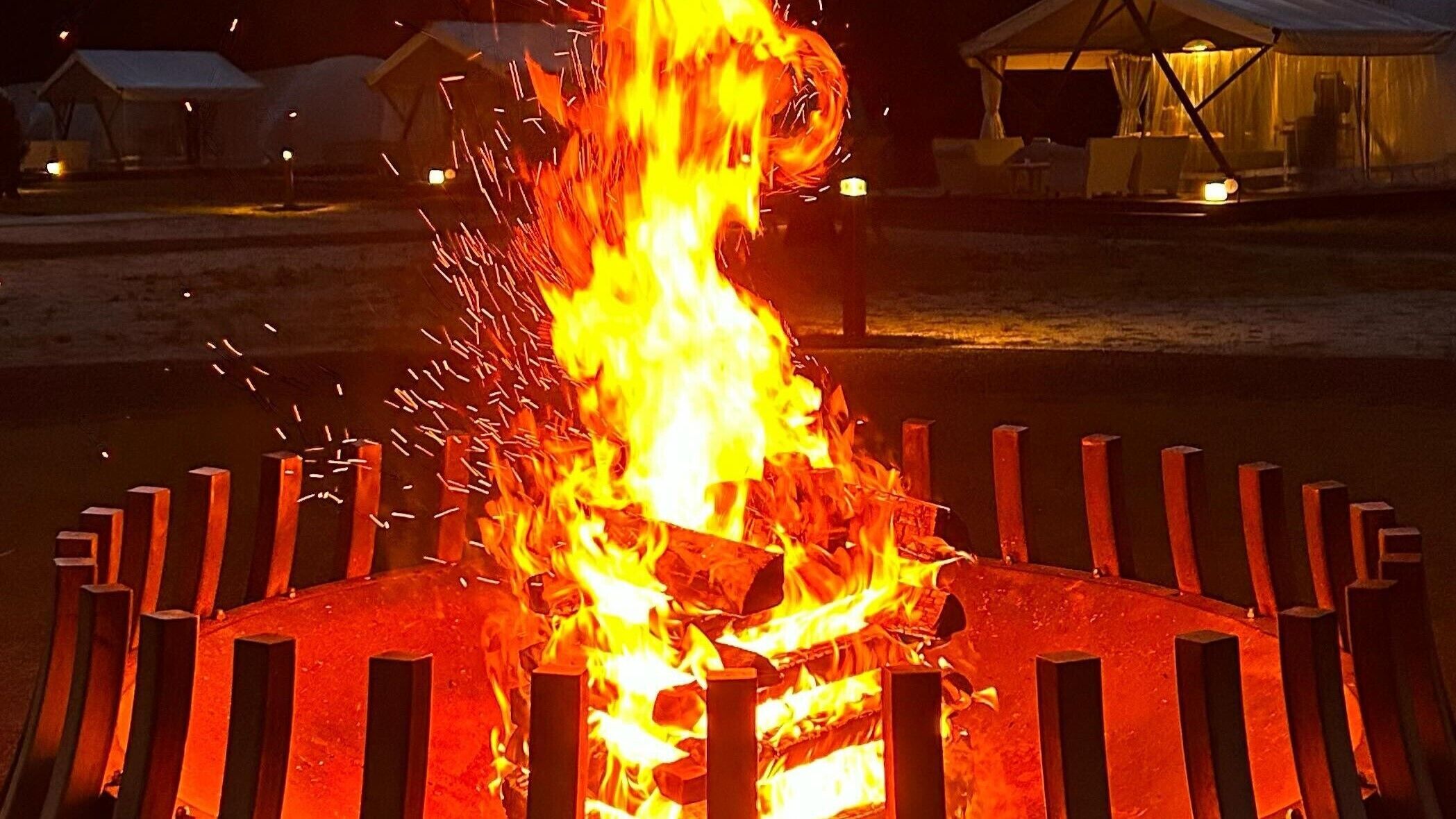 A campfire with a metal fence surrounding it, set in an outdoor area with tents in the background.