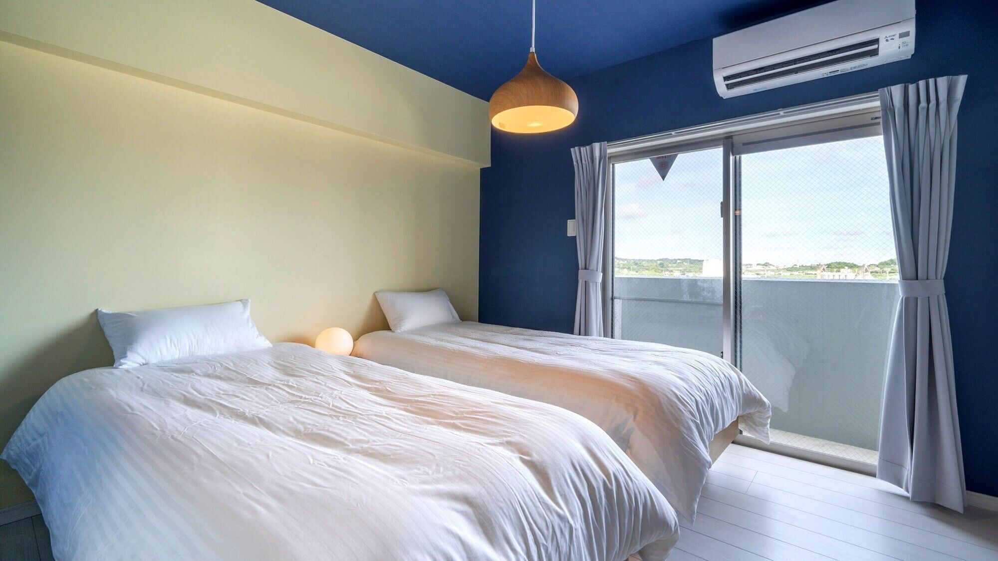 A bedroom with two beds, a hanging light fixture, an air conditioning unit, and a view of the outdoors through a sliding glass door.
