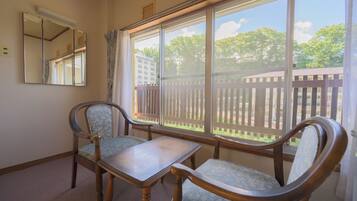 A room with a wooden table and two chairs, a large window with a view of a fence and trees, and a mirror on the wall.