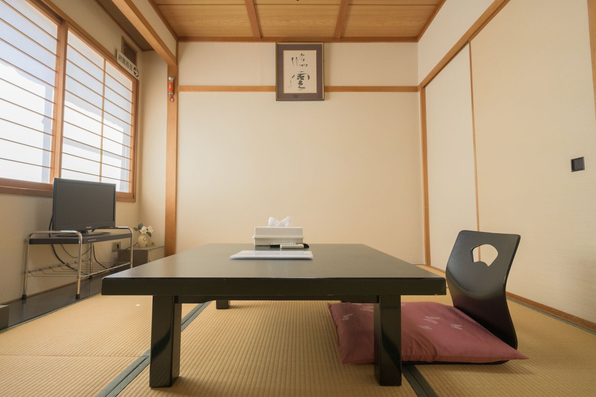 A tatami mat room with a low table, a chair, a television, and a framed calligraphy piece on the wall.