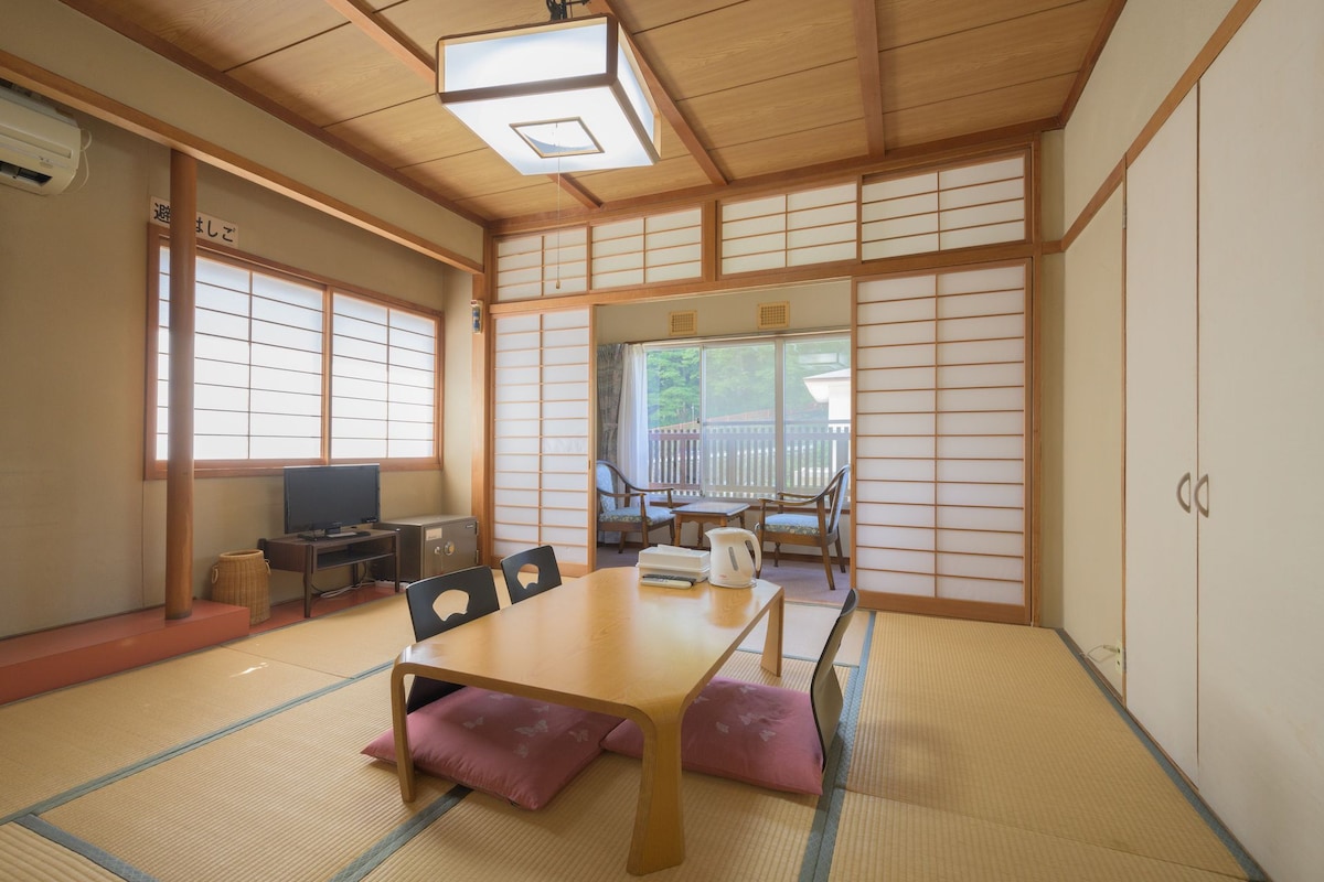 Um quarto japonês tradicional com tatames, uma mesa baixa, cadeiras, uma televisão e portas deslizantes.