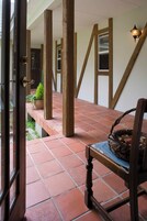 Uma porta aberta dá acesso a um alpendre coberto, com vigas de madeira, piso ladrilhado e uma cadeira com cesta.