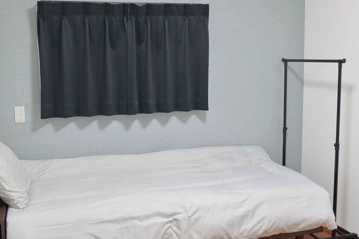 Een kleine eenpersoonskamer met een eenvoudig bed, een donker gordijn en een simpele metalen standaard.