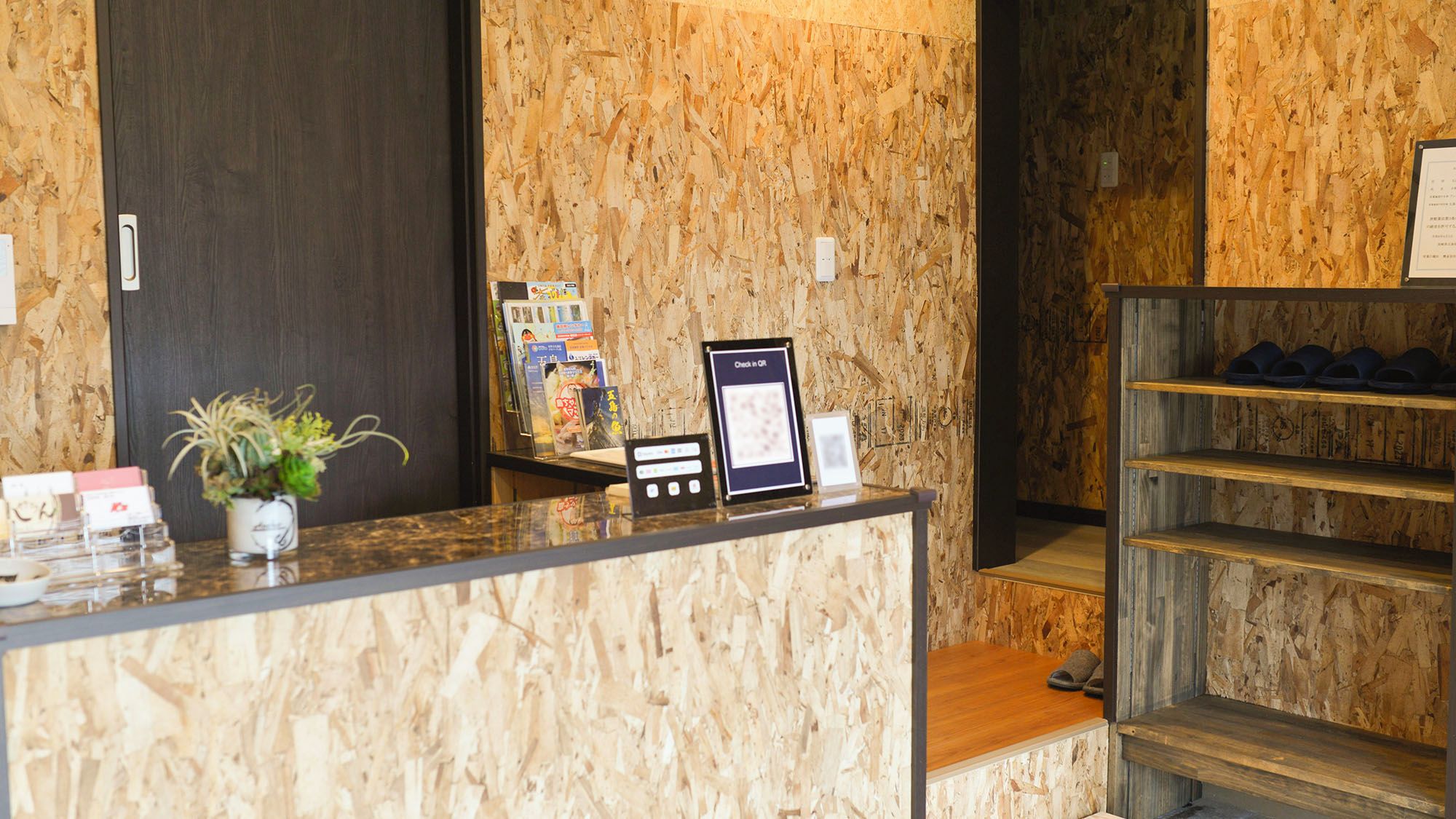 Una recepción con fondo de madera, una planta, un perchero para revistas y un cartel enmarcado.