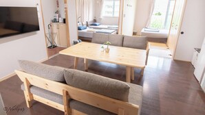 A modern, well-lit room with a wooden dining table and benches, a TV mounted on the wall, and a window with curtains. - Icony (Miyakojima)
