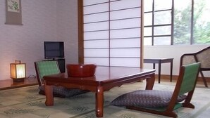 A traditional Japanese room with tatami flooring, a low wooden table, green cushioned seating, a television, and a window with a view of greenery. - Merci 0823 (Ito)