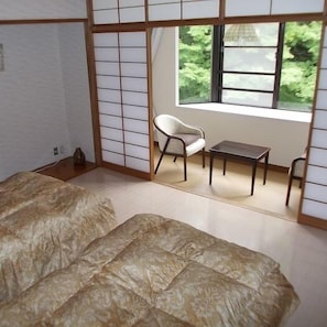 A traditional Japanese-style room with tatami flooring, a low table, a chair, and a view of greenery through a window. - Merci 0823 (Ito)