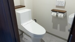 Inodoro blanco con un estante de madera encima, un dispensador de papel higiénico en la pared y un registro de fontanería a la vista.