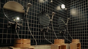 A modern bathroom with black tiled walls, multiple showerheads, and wooden stools. - EN+ Sauna & Capsule Bed (Kochi)