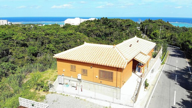 Um pequeno edifício térreo com telhado de cerâmica, cercado por vegetação densa, situado perto de uma estrada com vista para o mar.