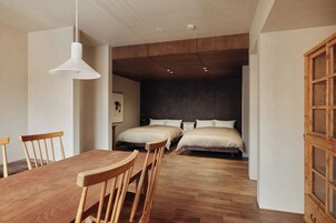 Una habitación moderna con una mesa de comedor de madera, dos sillas de madera, una lámpara colgante blanca, una cama con ropa de cama blanca y un armario de madera.