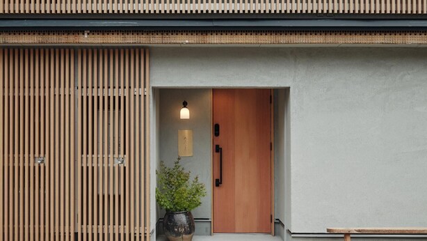 Een moderne entree van een gebouw met een houten deur, een plant in pot en een bankje buiten.