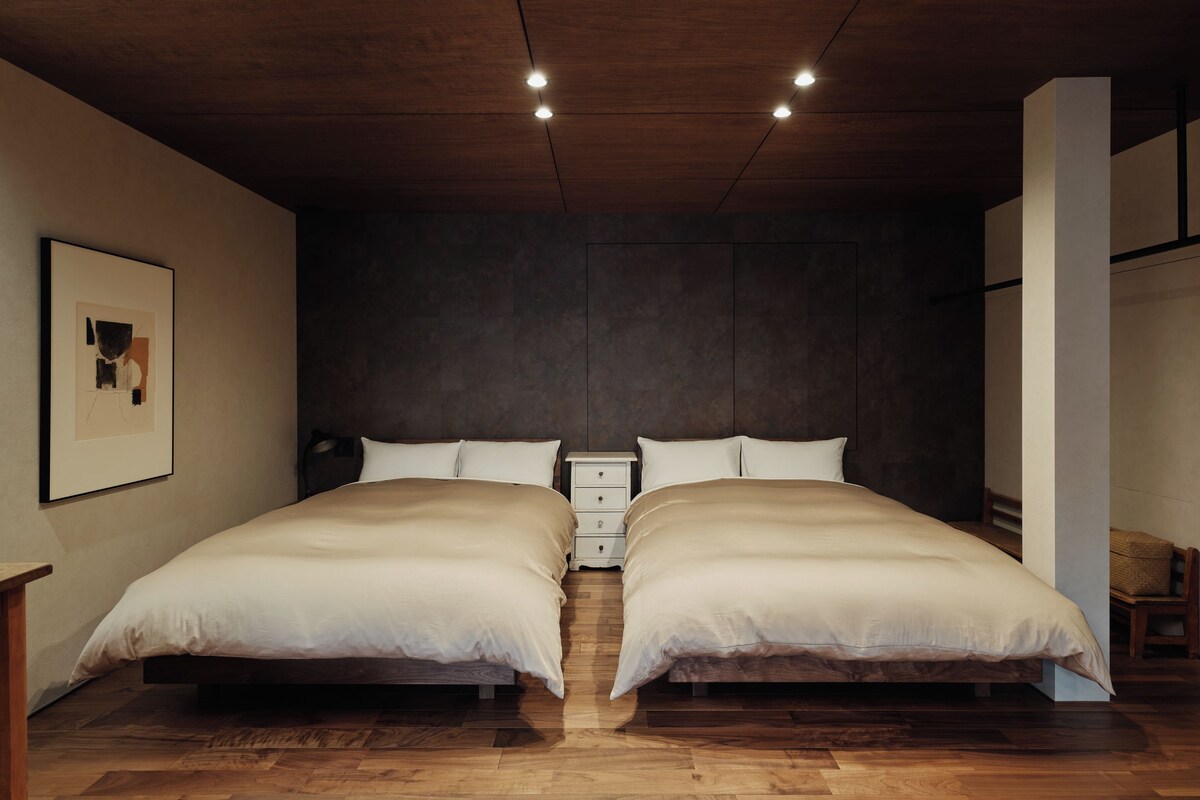 Une chambre avec deux lits, un plancher en bois, un mur de couleur sombre et un tableau encadré sur le côté gauche.