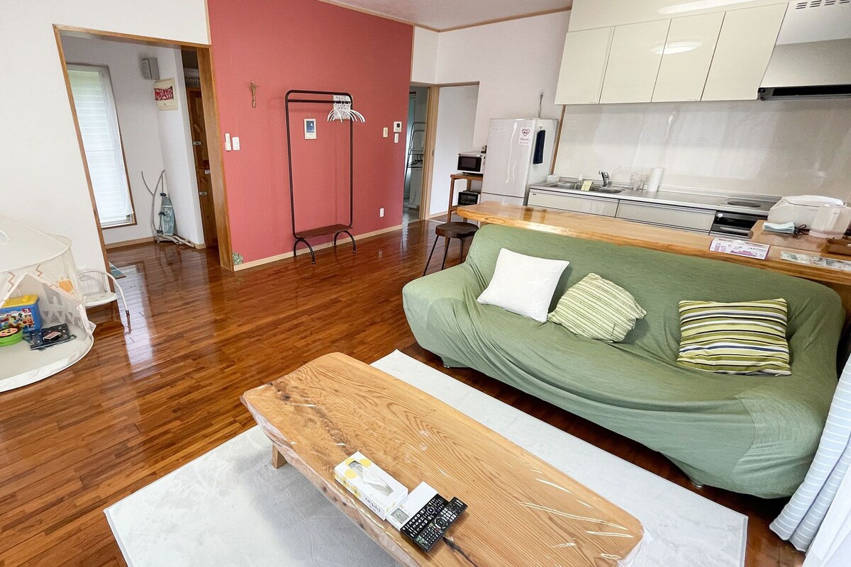 Un espacio habitable compacto con piso de madera, un sofá verde con almohadones, una mesita de centro blanca, un área de juegos tipo tienda de campaña blanca y una kitchenette con gabinetes y electrodomésticos blancos.