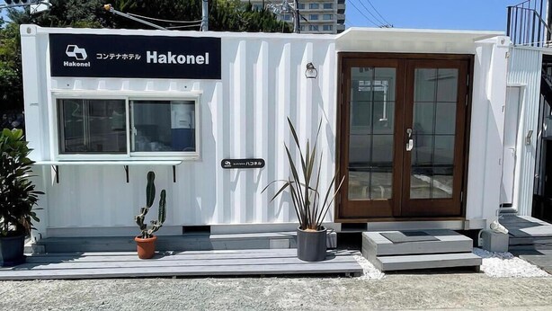Un edificio compacto y moderno con un letrero que dice "Hakonel". Tiene una puerta de madera grande, una ventana pequeña y una planta en maceta a nivel de calle.