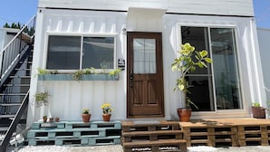 Un pequeño edificio blanco con puerta de madera, dos ventanas y un porche con plantas en macetas.
