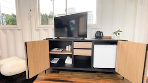Una habitación compacta y moderna con televisión integrada, un refrigerador blanco pequeño y un escritorio de madera con hervidor y una planta.