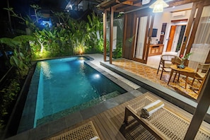 A swimming pool with a wooden deck, surrounded by lush greenery and a small seating area with a table and chairs.