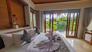 Habitación de hotel con cama, almohadas, una mesita con una flor, una silla verde y vistas a un jardín a través de una puerta corrediza.