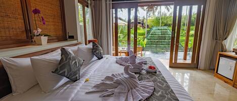 Habitación de hotel con cama, almohadas, una mesita con una flor, una silla verde y vistas a un jardín a través de una puerta corrediza.
