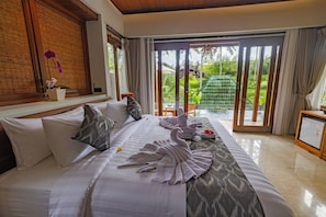 A hotel room with a bed, pillows, a small table with a flower, a green chair, and a view of a garden through a sliding door.