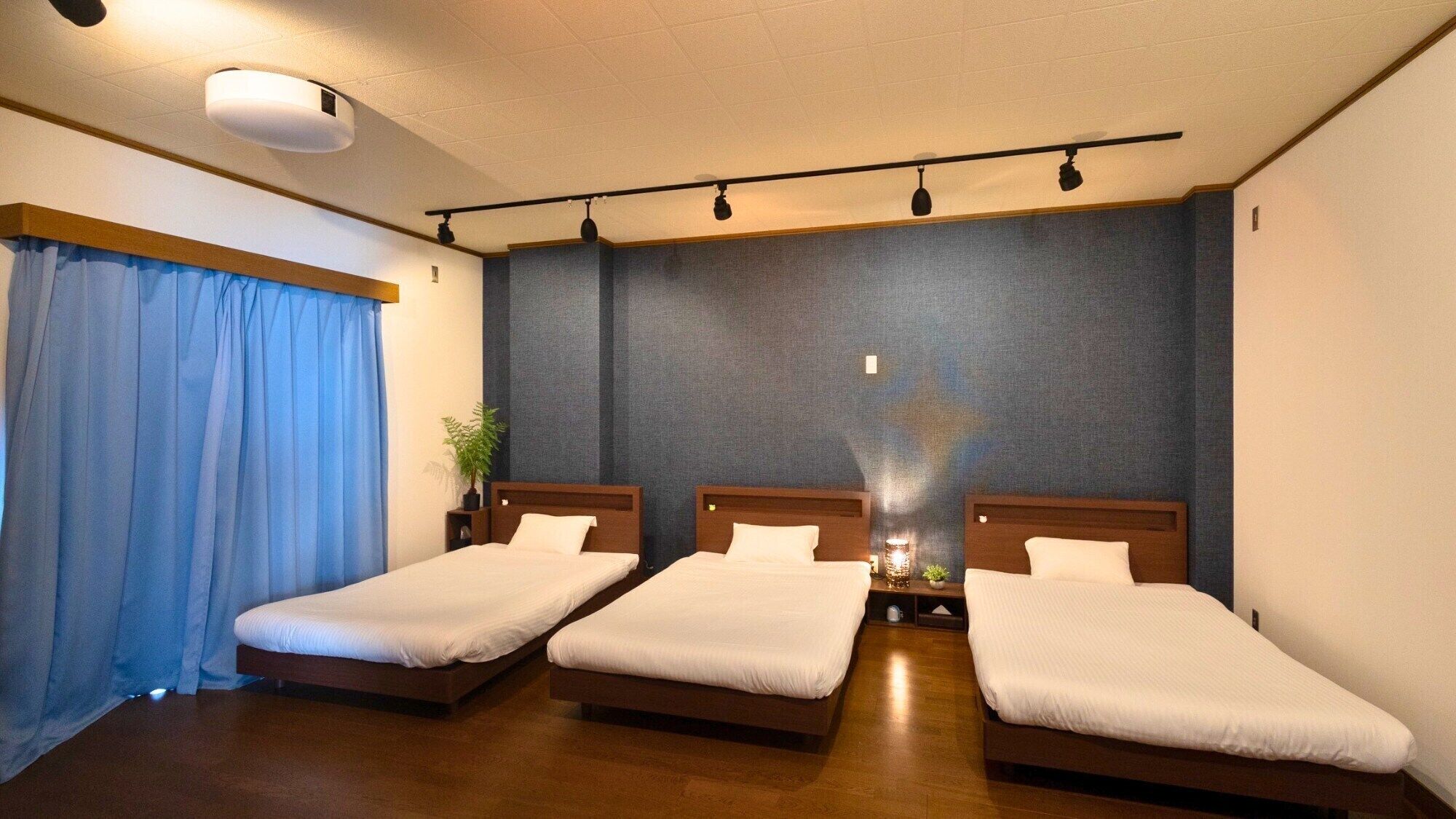 A hotel room with three single beds, a large window with blue curtains, a ceiling-mounted light fixture, and a wall-mounted shelf with a plant.