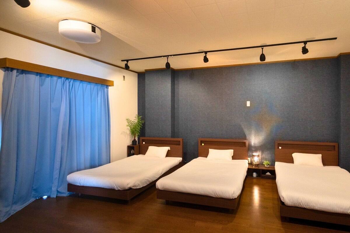 Una habitación de hotel con tres camas individuales, un ventanal grande con cortinas azules, una lámpara del techo y un estante en la pared con una planta.