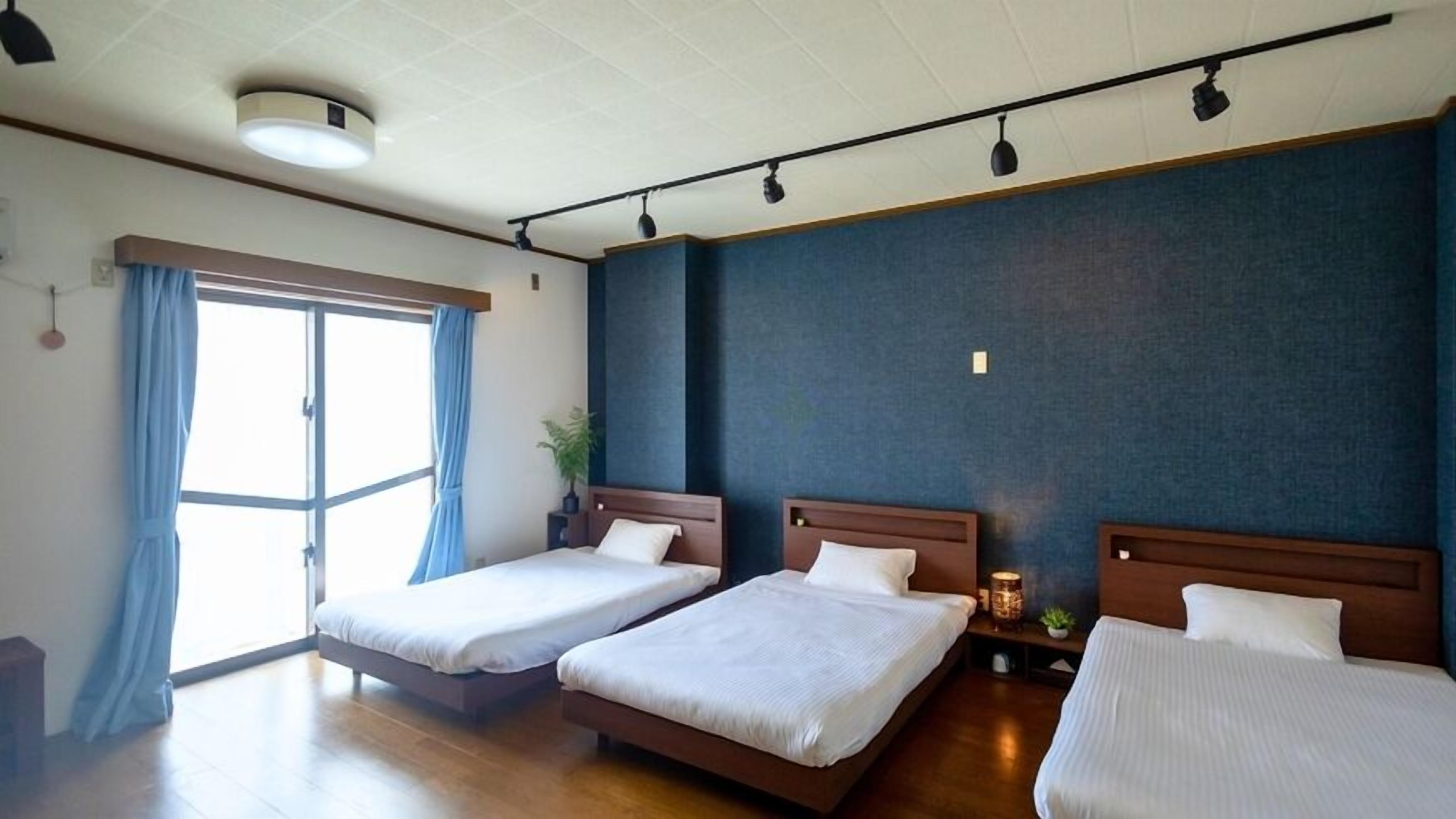 A hotel room with two beds, a large window with curtains, a ceiling light, and a wall-mounted lamp.