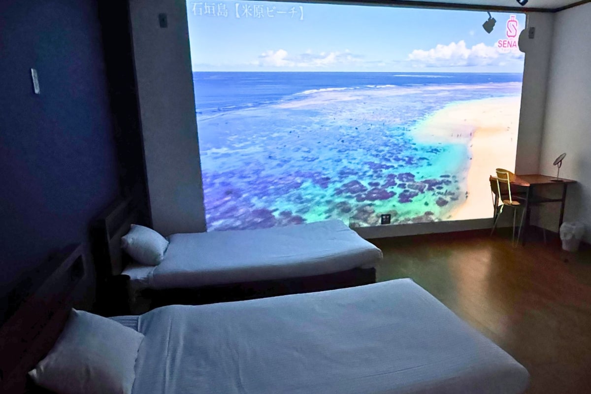 Quarto com duas camas, uma escrivaninha e uma janela grande com vista para a praia.