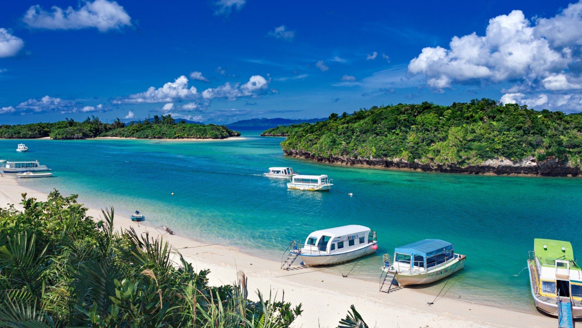 A beach with several boats docked, clear turquoise water, and lush greenery.