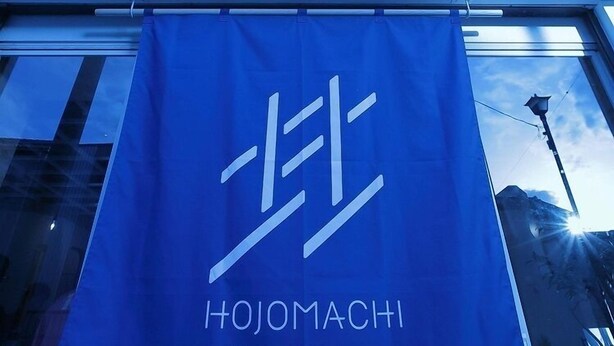 Une banderole bleue portant l’inscription « HOJOMACHI HOSTEL » est accrochée devant un immeuble.