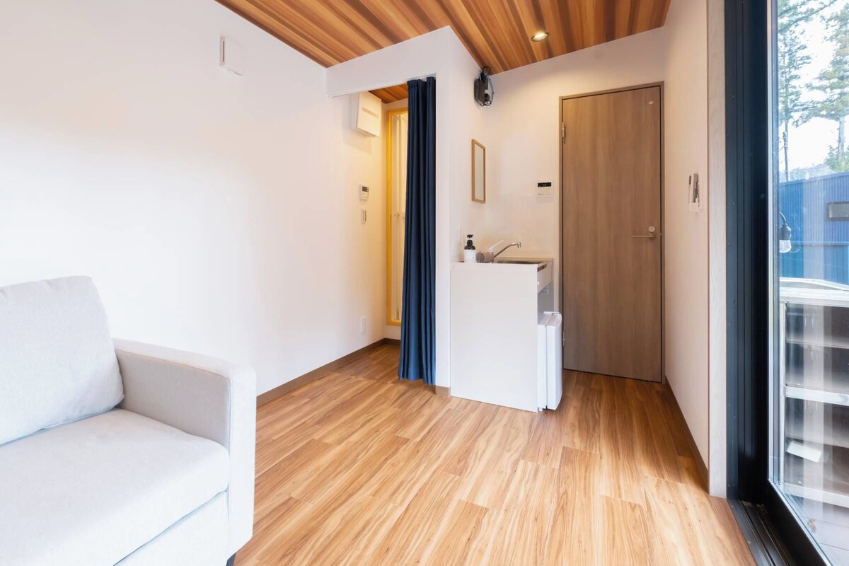 Ein modernes Zimmer mit Holzboden, einem weißen Sofa, einer Küchenzeile und einer Tür zu einem weiteren Bereich.