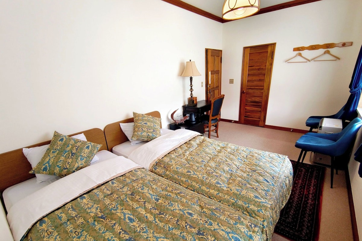 Quarto de hotel com uma cama grande, uma escrivaninha com cadeira, uma luminária e um guarda-roupa.