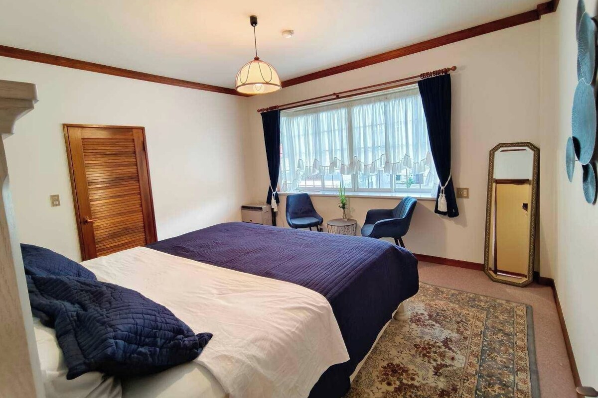 Quarto de hotel com cama, duas cadeiras, uma mesinha, janela com cortinas e espelho.