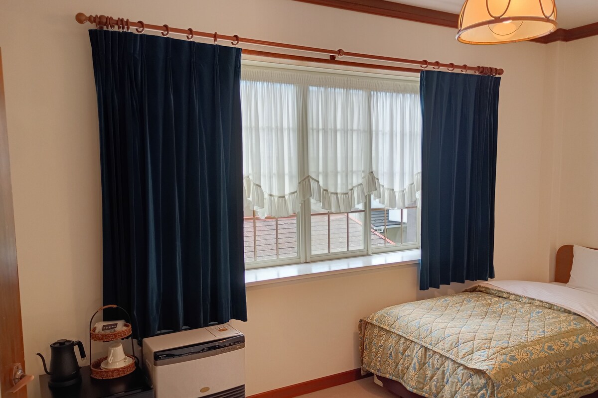 Quarto com cama, janela com cortinas, aquecedor e uma pequena mesa com chaleira.