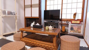 A traditional Japanese-style living room with tatami flooring, a low table with a woven top, wicker furniture, a television, and a small cabinet with decorative items.