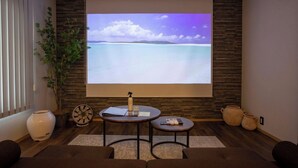 A room with a large screen displaying a scenic view, a small table with a bottle, a remote control, and a notebook.