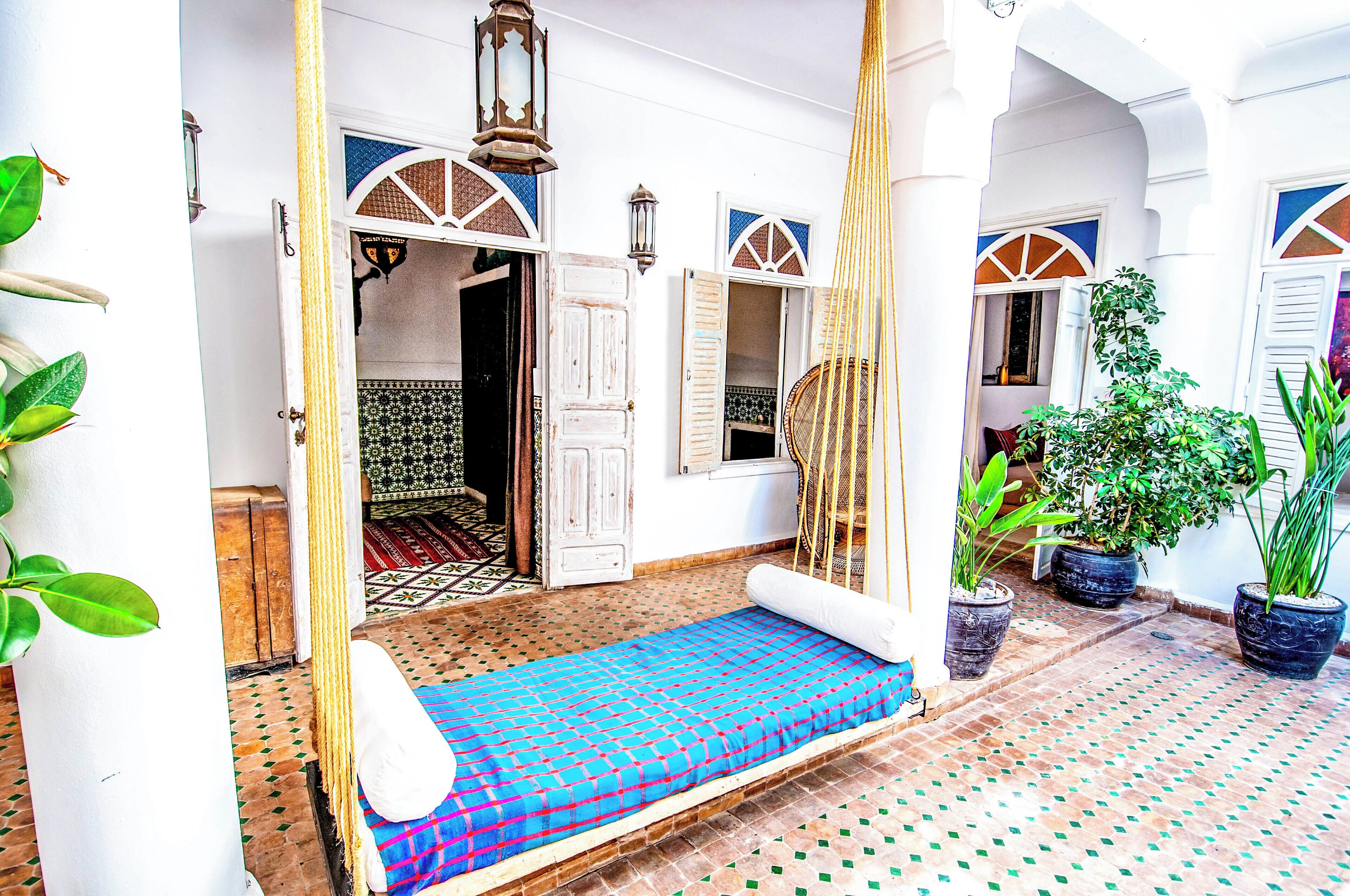 A traditional Moroccan room with a patterned floor, a bed with a blue and red bedspread, and potted plants.