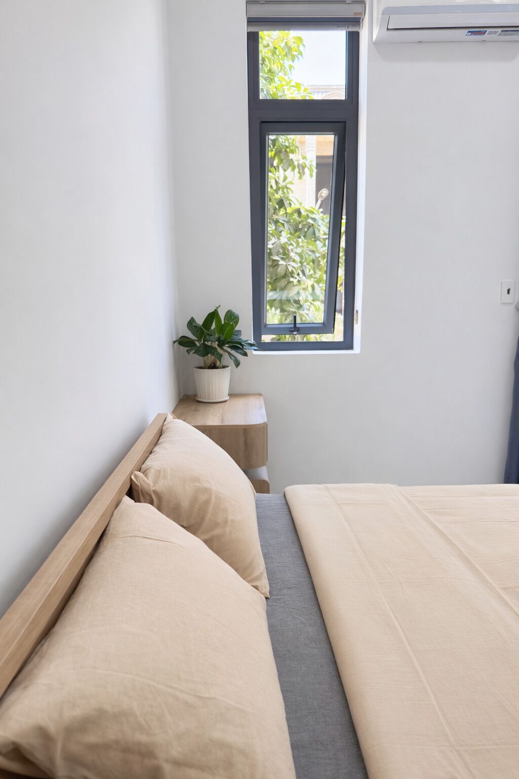 A bedroom with a bed, a nightstand with a plant, and a window with a view of greenery.