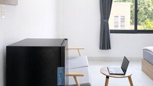 Una habitación moderna con un mueble negro, una silla de madera, una computadora portátil sobre una mesita y una ventana con cortina gris.