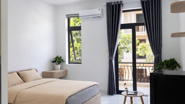 Un dormitorio moderno con una cama grande, una mesita con una computadora portátil, un equipo de aire acondicionado y un balcón con vistas a zonas verdes.