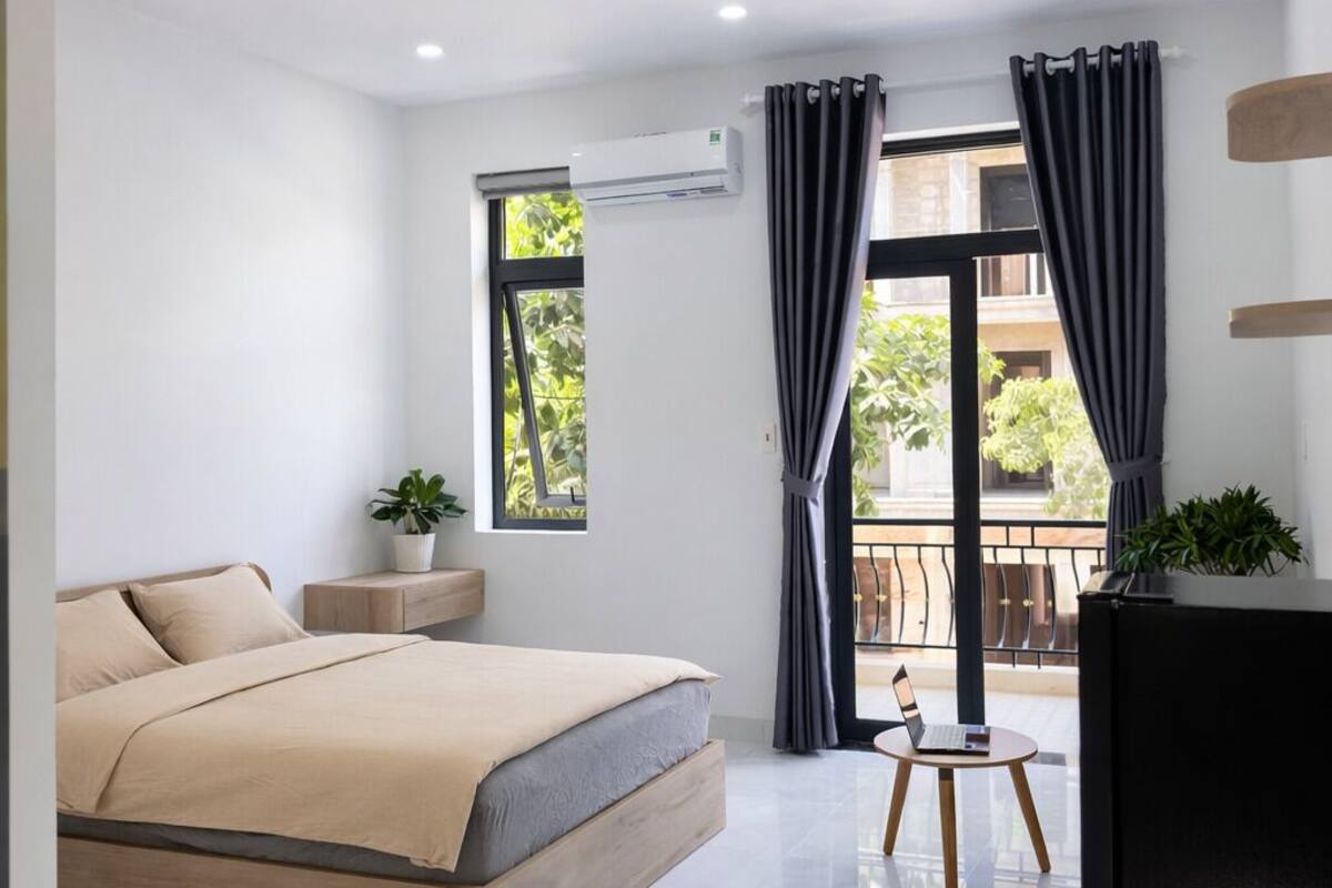 Un dormitorio moderno con una cama grande, una mesita con una computadora portátil, un equipo de aire acondicionado y un balcón con vistas a zonas verdes.