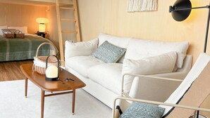 Ein Zimmer mit einem Etagenbett, einer Schlafcouch, einem Sessel, einem Tisch mit Lampe sowie einer Wandlampe.