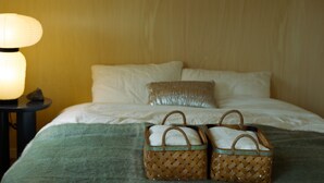 Ein Bett mit weißen Leinentüchern, einer grünen Tagesdecke und zwei Weidenkorb-Körben mit Tragegriffen.