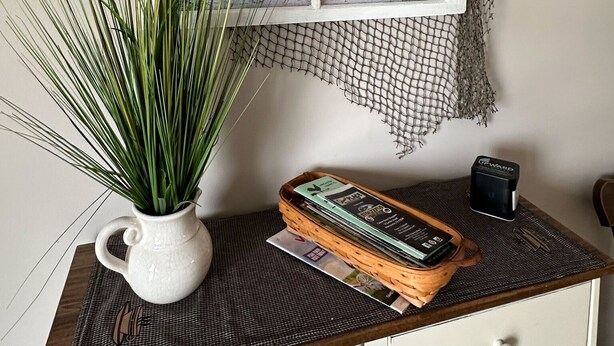 A framed painting of a beach scene with a window, a potted plant, and a stack of magazines on a wooden table. - Hilltop Guesthouse - in Intercourse on Beacon Hill, 4-Bedroom House (Ronks)