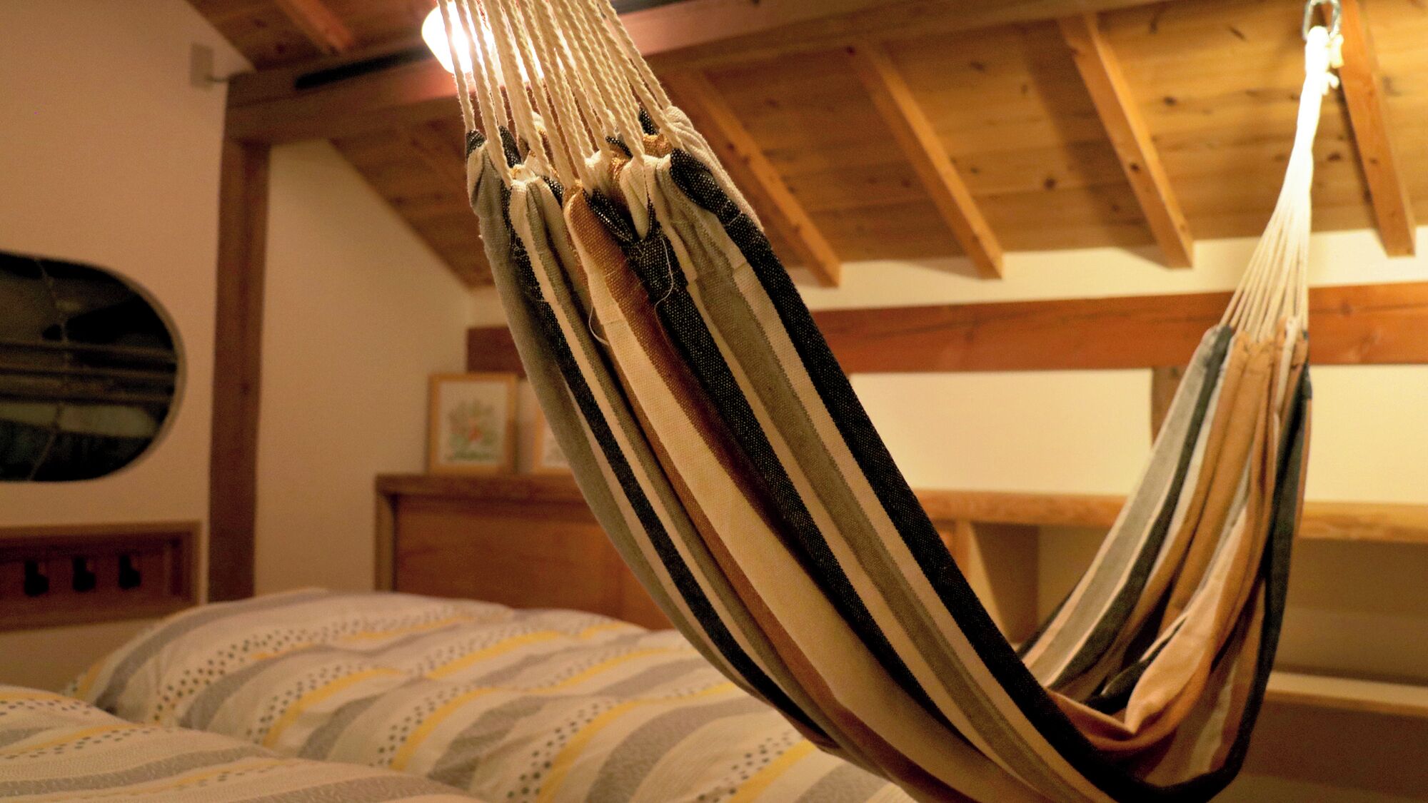 Uma rede em um quarto com vigas de madeira, uma cama com roupa de cama listrada e um quadro em cima de uma mesinha de cabeceira de madeira.